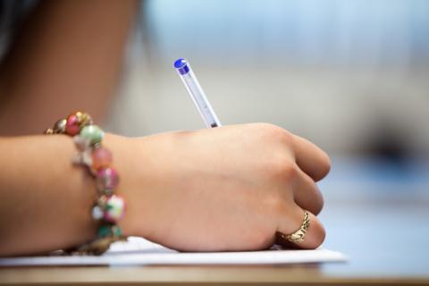 Lähikuva nuoren henkilön kädestä kirjoittamassa kuulakärkikynällä. Nuoren ranteessa on värikäs rannekoru.