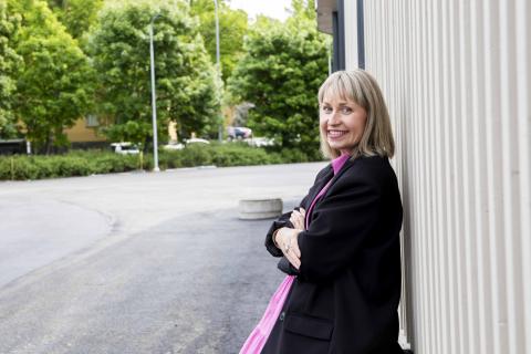 Henkilö nojaa seinään ja hymyilee. Taustalla on puita.