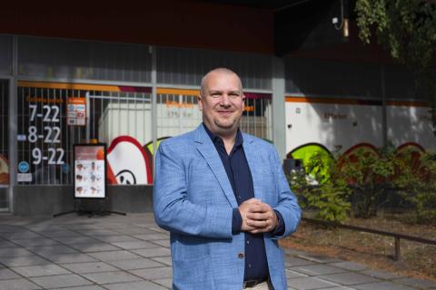 Henkilö seisoo ulkona lähikaupan edessä ja hymyilee.