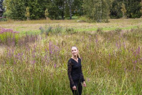 A stormwater expert, wearing dark clothes, standing in a green landscape. There are trees in the background.