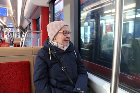 Eläkeikäinen rouva istuu ratikassa hymyillen ja katsoo ulos ikkunasta.