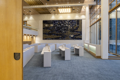 A hall with a sliding door that is slightly ajar. The hall has many small light-colored tables and a large table between the pillars. In the background is a large ceramic artwork.
