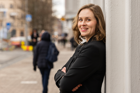 Hymyilevä nainen nojaa seinään tumma jakku yllään.