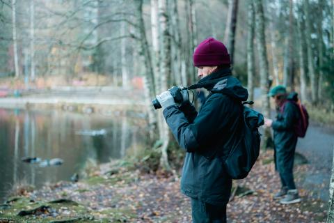 Valokuvaaja seisoo syksyisessä puistossa lammen rannalla. 