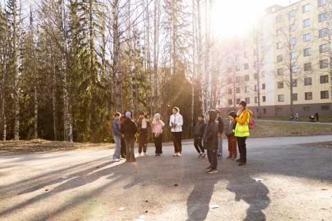 Kaksi opettajaa ja oppilaat seisovat puoliympyrässä metsän reunassa. Taustalla näkyy kerrostalo.